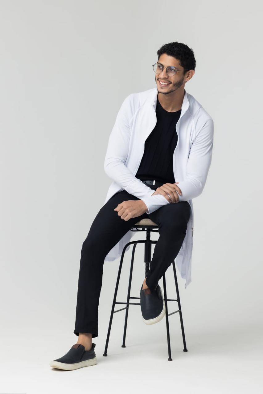 Dr. Davi Teixeira de jaleco branco, sentado em um banco alto, em retrato de corpo inteiro
